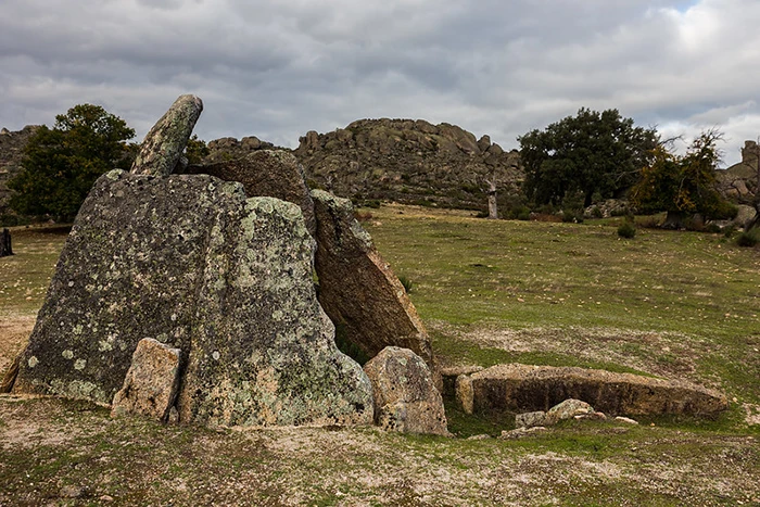Vista lateral del Dolmen Data I: ortostatos graníticos con líquenes y corredor semienterrado; berrocal de La Data y encinas al fondo en un día nublado.