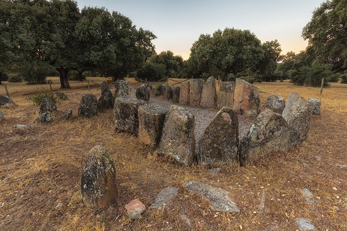 Vista frontal del Dolmen de la Gran Encina al atardecer, con la cámara funeraria rodeada de ortostatos de granito y encinas al fondo.