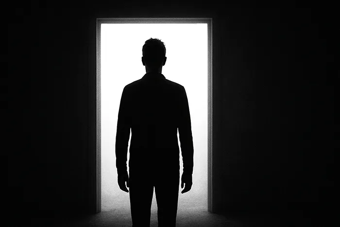 Silueta en blanco y negro de una persona de pie frente a un umbral iluminado, rodeada de oscuridad, representando el misterio y la fuerza del silencio.