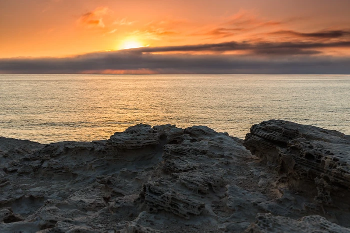 Roca volcánica oscura en primer plano con el sol asomando entre nubes bajas y cielo dorado.