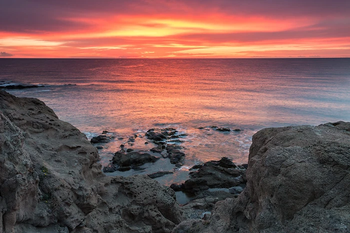 Amanecer rojizo entre peñascos, con el mar y las rocas recortadas en silueta.