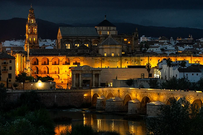 Vista nocturna de la Mezquita Catedral de Córdoba iluminada, con la torre y el puente romano reflejados en el río Guadalquivir.