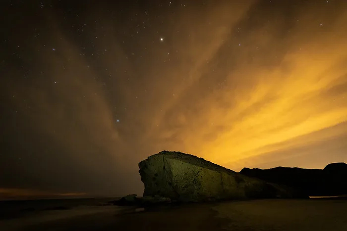 Playa de Mónsul de noche con nubes iluminadas y roca volcánica en primer plano.