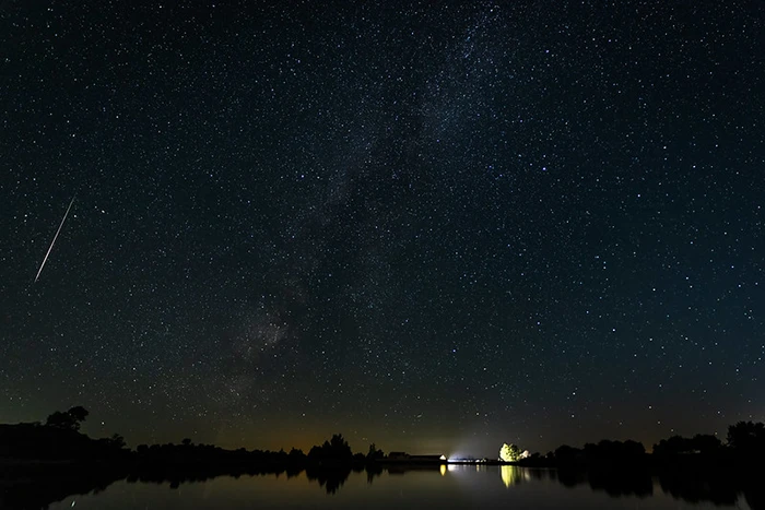 Vía Láctea y una estrella fugaz reflejadas en las aguas tranquilas de Los Barruecos con luces suaves en la distancia.