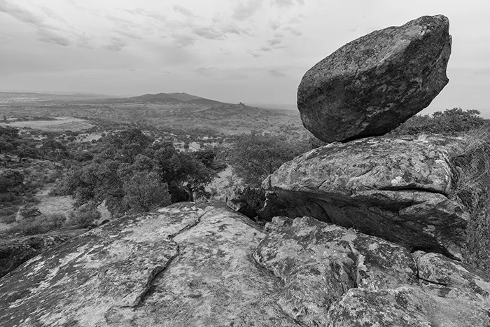 Bosque de encinas y alcornoques entre formaciones rocosas en Valcorchero, Plasencia, bajo un cielo parcialmente nublado en blanco y negro.
